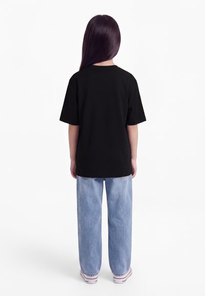 Enfant aux cheveux longs et foncés, de dos, portant un t-shirt noir, un jean bleu clair et des baskets blanches sur un fond blanc.