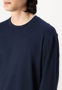 Tumši zils garām piedurknēm T-krekls, kas izgatavots no mīksta kokvilnas. Piedāvā kreiso kakla izgriezumu un diskrētu logotipa izšuvumu uz krūtīm. Gluda tekstūra.