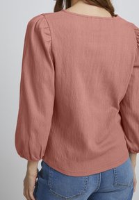Top de manga larga en rosa empolvado con hombros plisados, tejido texturizado y escote redondo, combinado con jeans de mezclilla azul claro.