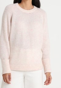 Weicher, hellrosa, fuzzy Strickpullover mit gerippten Bündchen und Saum, getragen mit weißen Hosen, aus der Schulter- bis Hüfthöhe gezeigt.