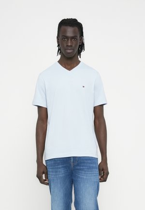 Mand med flettet hår iført en lyseblå V-hals T-shirt og blå jeans, stående foran en ensfarvet hvid baggrund.