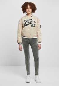 Beige satiinist bomber jope musta logo ja number 05, must-valged ribatud varrukad, koos hallide kitsaste teksade ja valgete tossudega.