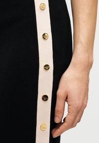 Μαύρο πλεκτό φόρεμα με αντίθετη ροζ πλαϊνή φάσα και έξι χρυσά κουμπιά κατά μήκος της πλευράς. Κοντινό πλάνο χεριού κοντά στα κουμπιά.