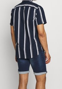 Mörkblå kortärmad skjorta med vita vertikala ränder, knappkrage och en avslappnad passform, parat med upprullade denimshorts.