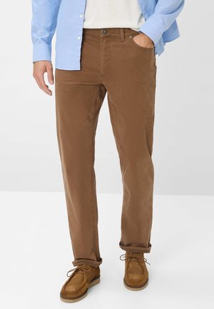 Braune Baumwollhose mit geradem Schnitt, vorderen Taschen und hochgekrempelten Säumen, kombiniert mit braunen Wildlederschuhen und einem hellblauen Hemd mit Knöpfen.