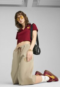 Camiseta polo corta roja con logo, pantalones jogger beiges con cordón, bolso cruzado negro, gafas de sol amarillas y zapatillas burdeos con detalles marrones.