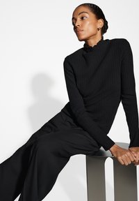 Haut noir à manches longues côtelé avec un col montant et une bordure festonnée, associé à un pantalon noir ample, assis sur un tabouret géométrique.