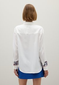 Camicia bianca a maniche lunghe con motivi paisley ricamati in rosso e blu sui polsini; tessuto liscio e vestibilità comoda.