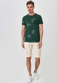 Grün gestreites T-Shirt mit weißen Schriftzug, runder Ausschnitt, kombiniert mit beigefarbenen Cargo-Shorts und weißen Turnschuhen. Einfaches, lässiges Design.