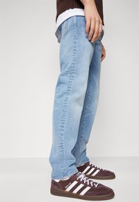 Blugi din denim bleu deschis cu o croială dreaptă și cusături vizibile, asortați cu adidași din nubuc violet închis, cu dungi albe.
