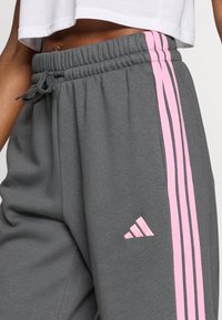 Szare spodnie dresowe z elastycznym pasem, różowymi paskami po bokach oraz różowym logo Adidas na dolnej części nogawki. Miękka tekstura materiału.