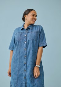 Femme aux cheveux foncés en chignon, portant une robe en denim bleu boutonnée à manches courtes, souriante et regardant sur le côté sur un fond uni.