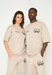 Camisetas oversized beige con logotipos bordados en negro, combinadas con pantalones beige a juego que presentan logotipos similares. Material de algodón suave.