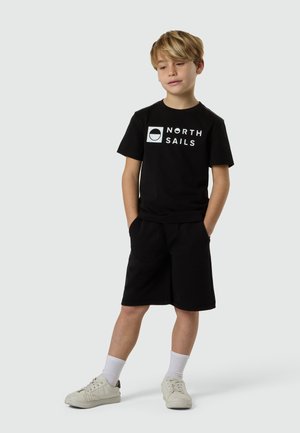 Garçon portant un t-shirt noir "North Sails", un short noir, des chaussettes blanches et des baskets blanches, debout les mains dans les poches contre un fond clair.