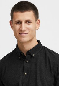 Camicia nera con bottoni, realizzata in tessuto testurizzato, caratterizzata da un colletto alla francese e bottoni tondi marroni. Capelli corti e indossa un piccolo orecchino a cerchio.