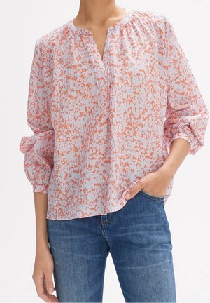 Frau trägt eine helle, langärmelige Bluse mit orangefarbenem abstraktem Muster, teilweise in blaue Jeans gesteckt, eine Hand in der Tasche.