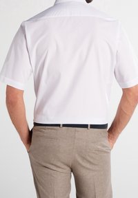 Camisa de vestir blanca de manga corta, fabricada con un tejido suave, combinada con pantalones color marrón claro que tienen un cinturón oscuro, vista desde la parte trasera.