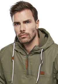 Veste à capuche vert olive en matière texturée, avec une fermeture éclair avant quart, des cordons blancs et une petite étiquette en cuir.