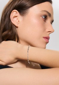 Srebrna bransoletka z przezroczystymi kamieniami, charakteryzująca się delikatnym wzorem łańcuszka i regulowaną długością, spoczywająca na jasnej karnacji skóry.