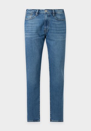Mand iført en lyserød T-shirt med korte ærmer med små tekst og symboler, blå jeans, brun bælte og gule loafers, stående med den ene hånd i lommen.