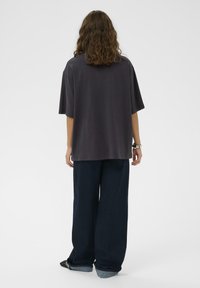 Vállig érő, göndör hajú személy, aki laza sötétszürke pólót, bő sötétkék farmert és mintás cipőt visel, háttal áll.