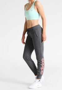 Lichtblauwe sportbeha, grijze joggerbroeken met roze 'adidas' logo. Materiaal lijkt zacht en elastisch; nauwsluitende pasvorm met taps toelopende enkel. Witte sneakers.