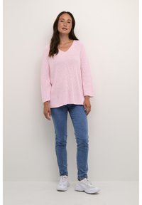 Růžový pletený svetr s výstřihem do V, doplněný modrými skinny džíny a bílými teniskami. Dlouhé rukávy a volný střih vytvářejí ležérní vzhled.