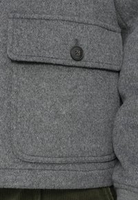 Tascone in lana grigia con un bottone nero etichettato "Hackett London" su un tessuto di velluto a coste verde.