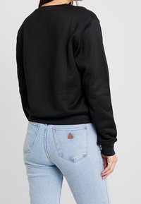 Felpa nera con scollo rotondo, polsini e orlo a costine. Abbinata a jeans azzurri chiari con un accento a triangolo in pelle marrone sulla tasca posteriore.