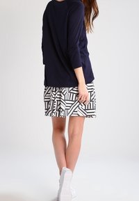 Woman standing with back turned, wearing a navy long-sleeve top, black and white geometric patterned skirt, and white sneakers.