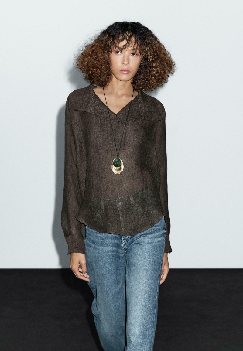 Brown, textured long-sleeve blouse with a v-neck and a peplum hem, paired with light blue jeans. Accompanied by a pendant necklace.