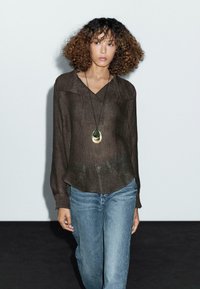 Blouse longue texturée brune à manches longues avec un décolleté en V et un ourlet peplum, associée à un jean bleu clair. Accompagnée d'un collier pendentif.