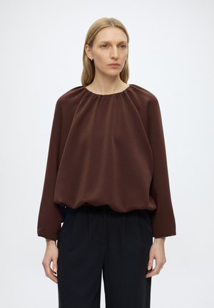 Femme aux cheveux blonds raides porte un chemisier marron ample à manches longues et un pantalon noir taille haute sur un fond uni.