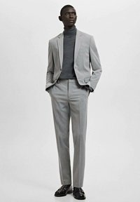 Man staat met handen in de zakken, gekleed in een lichtgrijs pak, donkergrijze coltrui en zwarte nette schoenen, tegen een effen achtergrond.