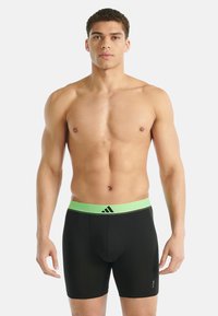 Atletický muž stojící čelem vpřed, oblečený v černých přiléhavých šortkách se zeleným páskem a logem Adidas na přední straně.