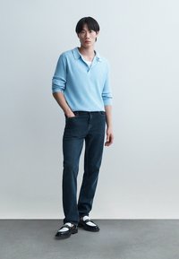 Mann in hellblauem, geripptem Langarmshirt, dunklen Jeans und schwarz-weißen Mokassins, der mit einer Hand in der Tasche vor einfarbigem Hintergrund steht.