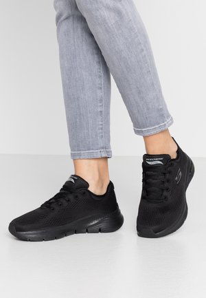 Zwarte sportieve schoenen van ademend mesh met veters en een gestructureerde zool. Ken features een S-logo aan de zijkant en "Arch Fit" branding op de hiel.