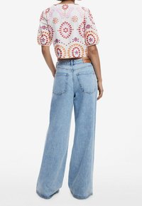Kvinde i lyseblå viddejeans og en hvid kortærmet bluse med røde, orange og lilla cirkulære mønstre, set fra ryggen.