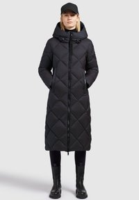Black quilted long coat with a hood, front zipper, and side pockets, paired with black leggings and ankle boots. Smooth, insulated texture.