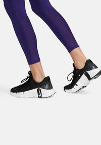 Piernas en leggings morados, usando zapatillas Nike en negro y blanco, un pie apoyado en el suelo y el otro ligeramente elevado sobre un fondo blanco.