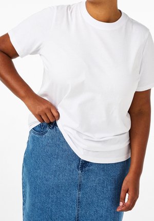 Persona que lleva una camiseta blanca lisa de manga corta metida ligeramente en una falda vaquera azul, de pie con una mano en el bolsillo.