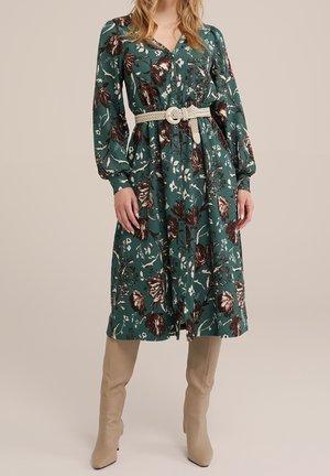 Frau trägt ein grünes, geblümtes Midikleid mit langen Ärmeln und einem gewebten beigen Gürtel, kombiniert mit kniehohen beigen Stiefeln.