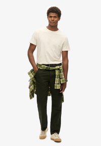Lichtbeige t-shirt, donkergroene broek en sneakers met een gestructureerde zool. Groene geruite blouse om de taille geknoopt. Eenvoudig ontwerp, casual pasvorm.