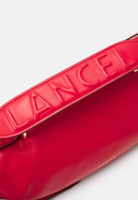 Sac à main en cuir rouge avec le nom de la marque en relief sur la bandoulière, design épuré, texture lisse et accents en métal doré.