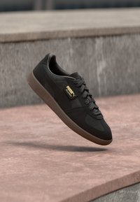 Crne tenisice Puma Palermo s gumenom petom lebde iznad ružičaste betonske površine i pozadine od sivog kamena.