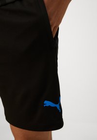 Bermudas deportivas negras con una textura suave. Presentan un logo de Puma azul en el lado izquierdo y un bolsillo lateral. Diseño simple y funcional.