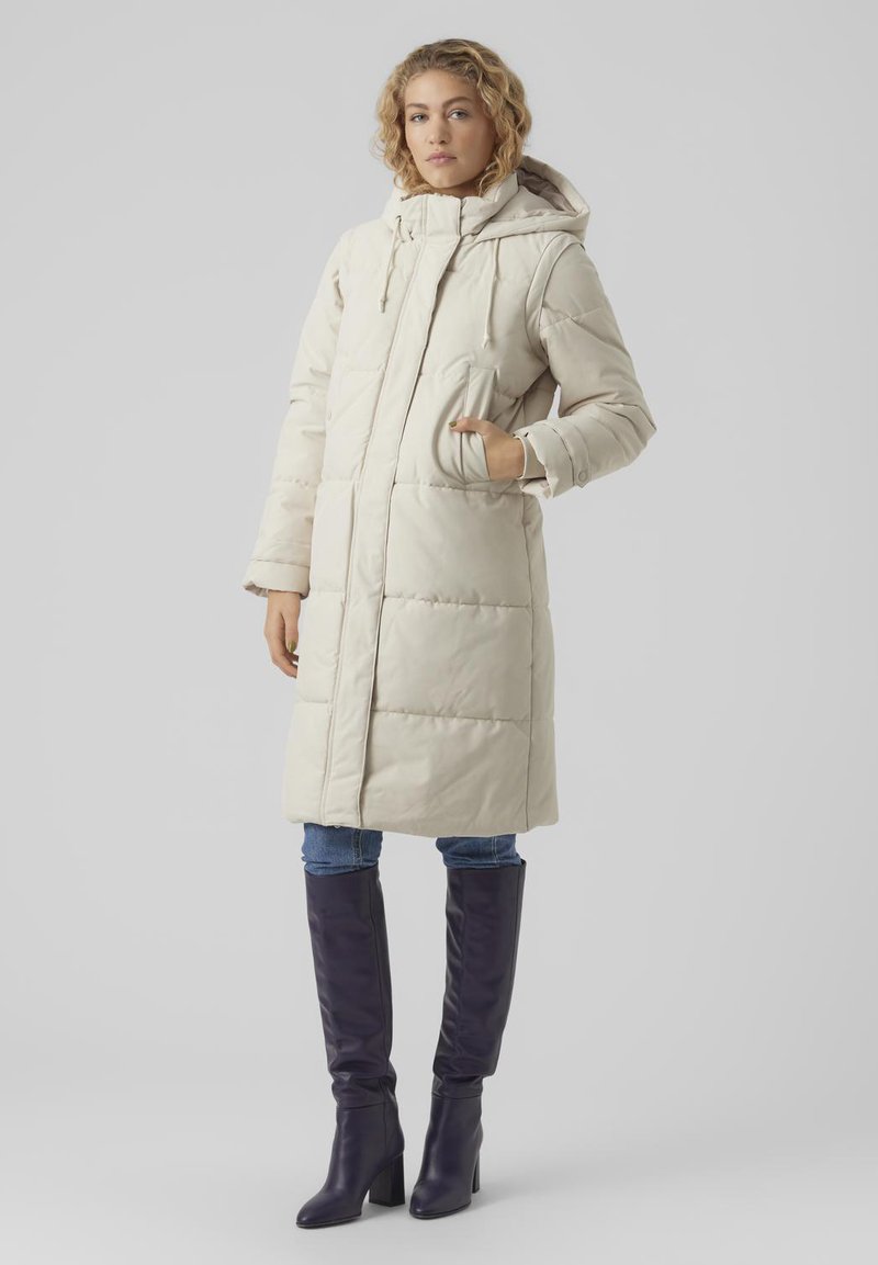 Lunga giacca puffer beige con cappuccio, dotata di chiusura con zip, tasche laterali e un motivo trapuntato. Abbinata a jeans blu e stivali viola al ginocchio.