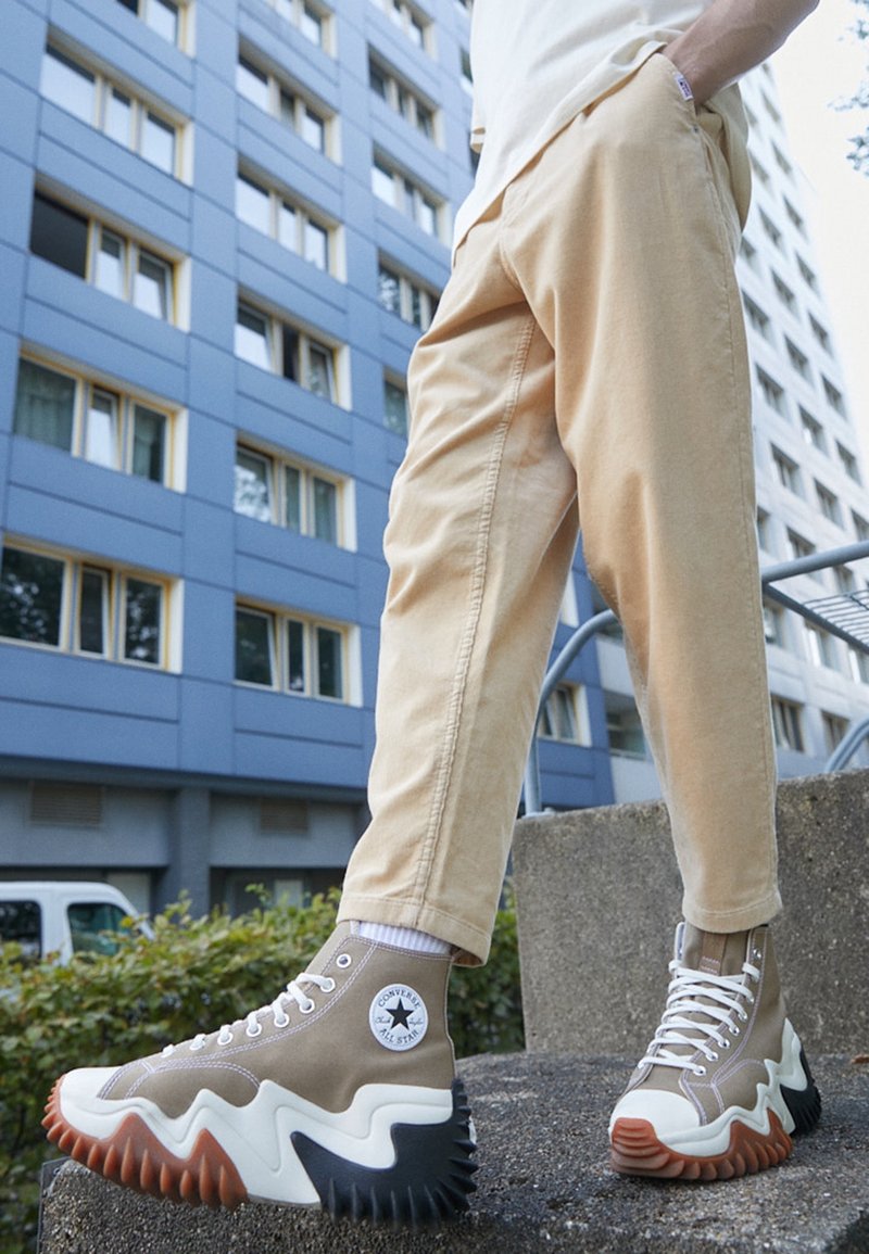 Personne portant des baskets montantes Converse beiges avec un dessin ondulé sur la semelle, debout sur des marches en béton devant un immeuble bleu.