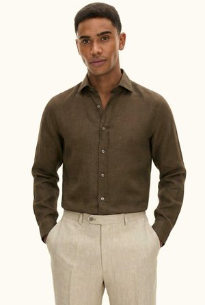 Mies, jolla on päällään tummanruskea nappikiinnitteinen pitkähihaisia paita ja vaaleanbeiget housut, kädet taskuissa, seisomassa yksivärisen taustan edessä.