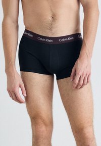 Črne bombažne boksarice z maroon pasom, na katerem je v belem napisana oznaka "Calvin Klein". Srednja višina zasnove z brezšivnim robom za udobje.
